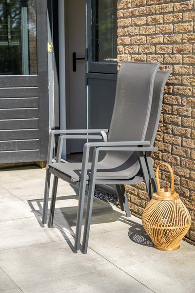 Zwei gestapelte graue Gartenstühle mit Armlehnen und Textilbespannung auf einer gefliesten Terrasse neben einer Backsteinwand und einer Laterne.