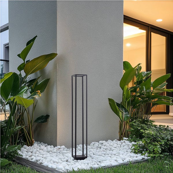 Moderne schwarze Sockelleuchte im offenen Rahmendesign in einem Beet aus weißen Steinen neben einer Hauswand und grünen Pflanzen.