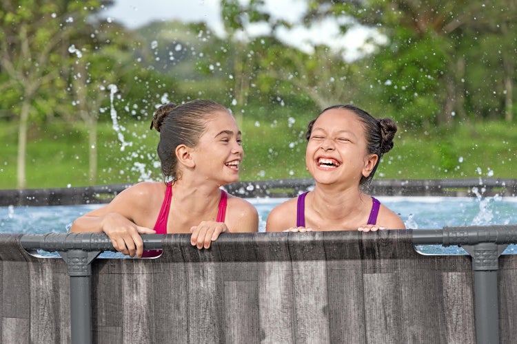 Zwei lachende Mädchen in einem Aufstellpool mit grauer Holzoptik und dunklem Rahmen, spritzendes Wasser im Garten.