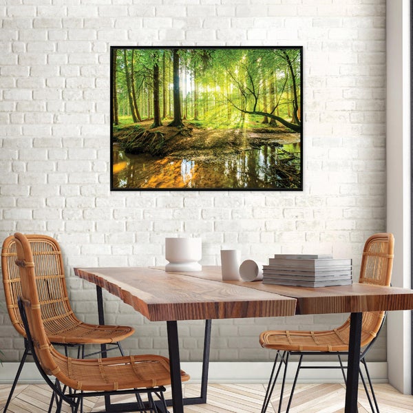Wandbild mit Waldmotiv und Bachlauf in schwarzem Rahmen über einem Holztisch mit Rattan-Stühlen vor einer weißen Ziegelwand.