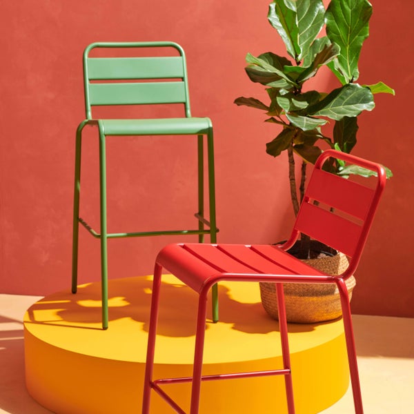 Ein grüner und ein roter Barhocker aus Metall mit Lamellen-Sitzfläche neben einer Zimmerpflanze in einem geflochtenen Korb.
