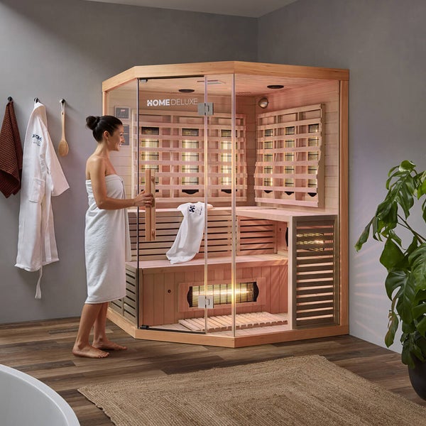 Eine Frau öffnet die Glastür einer Home Deluxe Eck-Infrarotkabine aus Holz, die in einem modernen Wellnessbereich mit Pflanze steht.