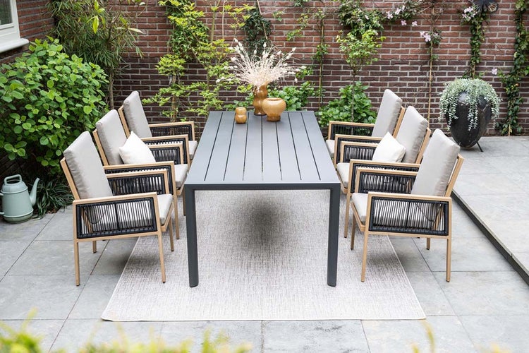 Moderne Gartensitzgruppe mit dunklem Metalltisch und sechs Holzstühlen mit Seilbespannung und Polstern auf einem Outdoorteppich auf einer Terrasse.