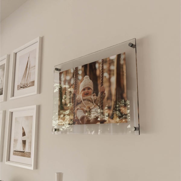 Ein rahmenloses Acrylglasbild mit einem Baby auf einer Schaukel, befestigt mit Wandabstandshaltern an einer hellen Wand neben gerahmten Bildern.