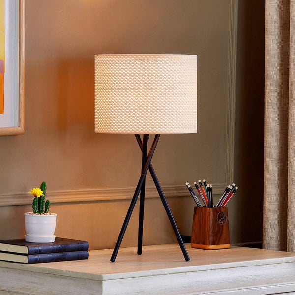 Tischleuchte mit dunklem Dreibein-Gestell und hellem, gemustertem Zylinderschirm auf einem Holzschreibtisch neben einem kleinen Kaktus.