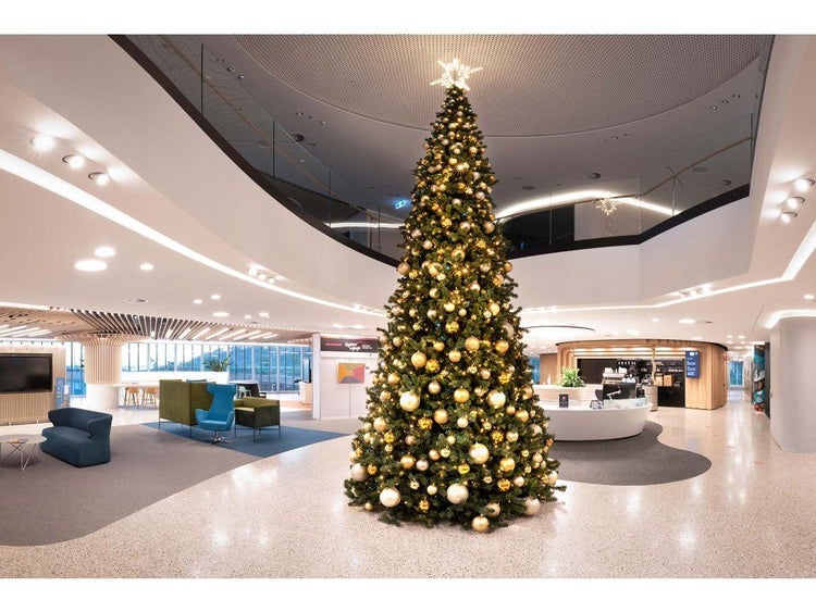 Ein großer festlich geschmückter Weihnachtsbaum mit Lichterkette und glänzenden Kugeln steht in einer modernen, weitläufigen Bürolobby.