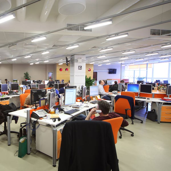 Helles, organisiertes Großraumbüro mit ergonomischen Arbeitsplätzen, Deckenbeleuchtung und orangefarbenen Bürostühlen.