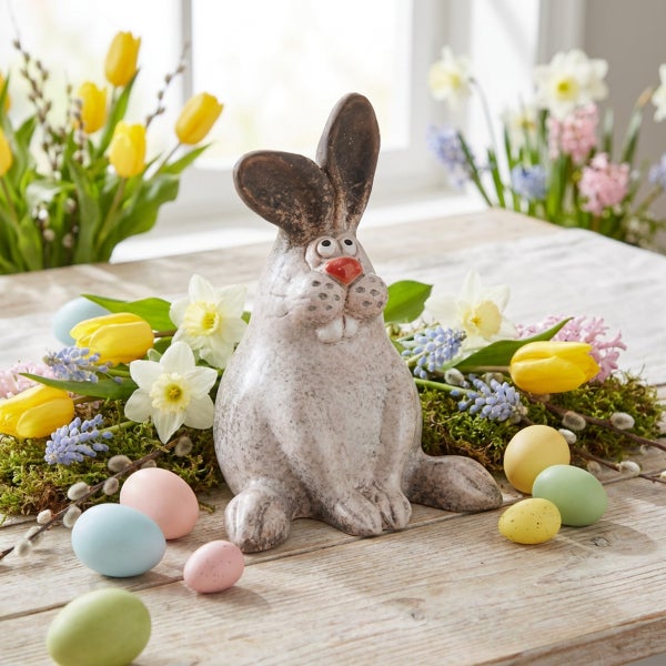 Osterhasenfigur aus Keramik als Tischdekoration umgeben von Frühlingsblumen und Ostereiern auf einem Holztisch.