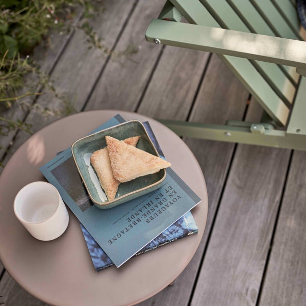 Kleiner runder Beistelltisch auf einer Holzterrasse mit einem Buch, Gebäck in einer Schale und einer Tasse neben einem grünen Holzgartenstuhl.