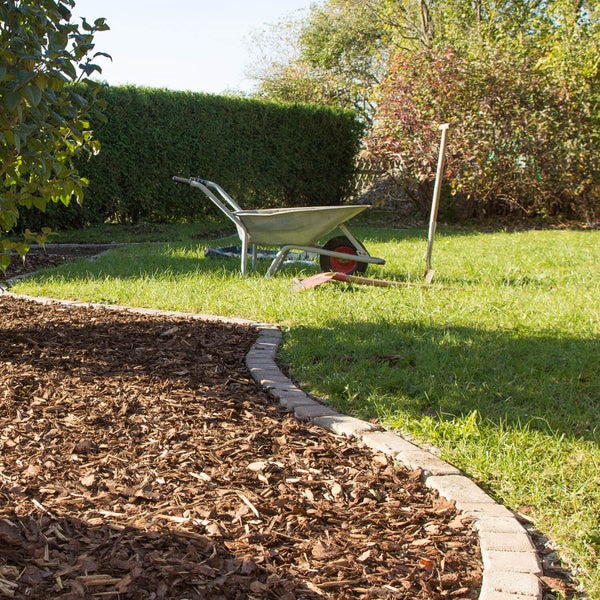 Gartenbeet mit Pinienmulch und Steinkante, im Hintergrund eine Rasenfläche mit Schubkarre und Schaufeln vor einer Hecke.