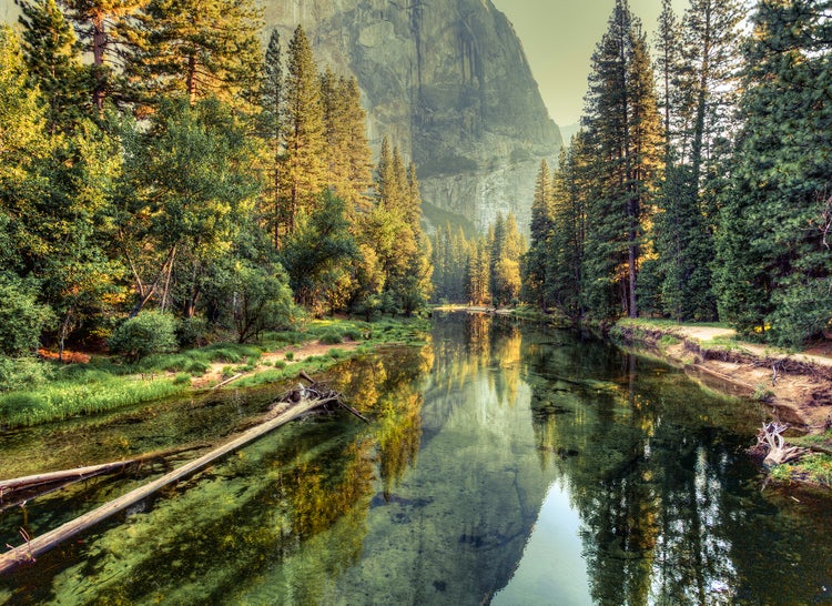 Landschaftsaufnahme eines ruhigen Flusses in einem Nadelwald mit einer großen Granitfelswand im Hintergrund und Spiegelungen auf dem Wasser.