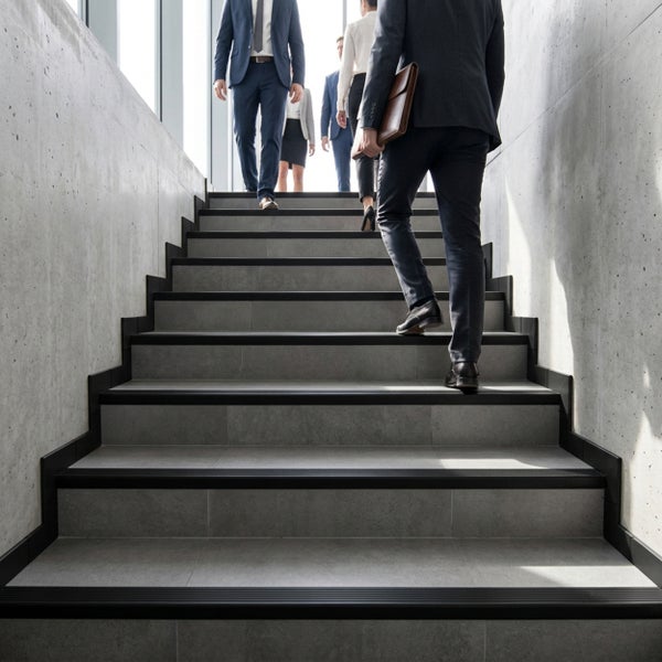 Moderne Treppe mit grauen Fliesen, dunklen Stufenkantenprofilen und Sichtbetonwänden, auf der Personen im Business-Outfit nach oben gehen.