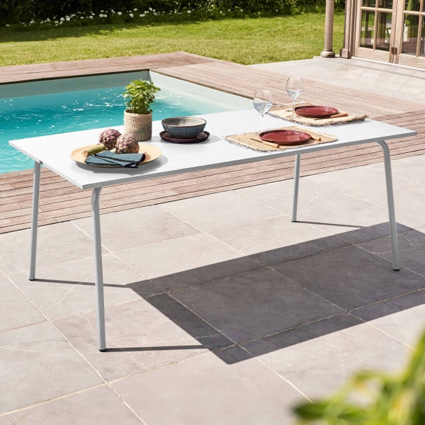 Weißer rechteckiger Gartentisch mit Metallbeinen auf einer Terrasse neben einem Pool, gedeckt mit Geschirr und einer Pflanze.