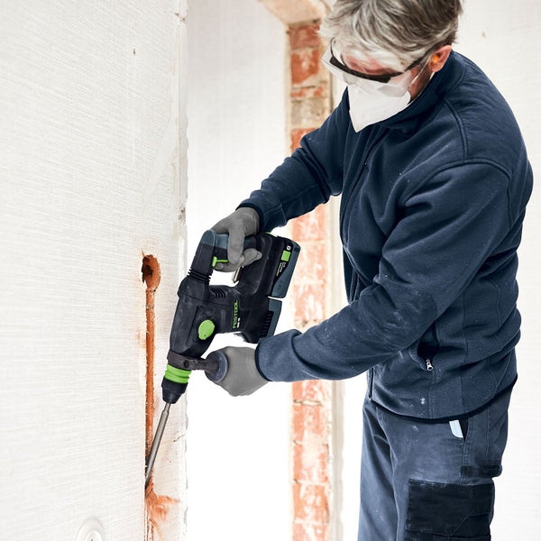 Handwerker bohrt mit einem Akku-Bohrhammer ein Loch in eine Wand. Er trägt dabei eine Schutzbrille und einen Atemschutz.