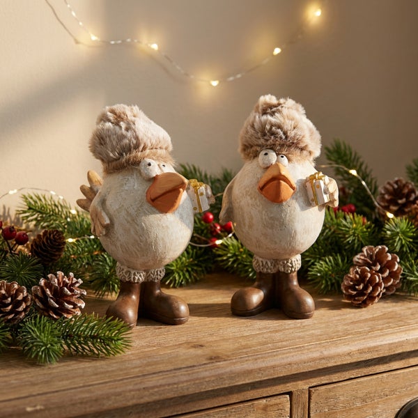 Zwei dekorative Hühnerfiguren als Winterdekoration mit Fellmützen und Stiefeln, gehalten auf einer Kommode mit Tannenzweigen und Lichterkette.
