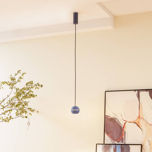 Moderne blaue Pendelleuchte mit kleinem kugelförmigen Schirm und langem Kabel in einem hellen, minimalistischen Raum mit Pflanze und Kunstwerk
