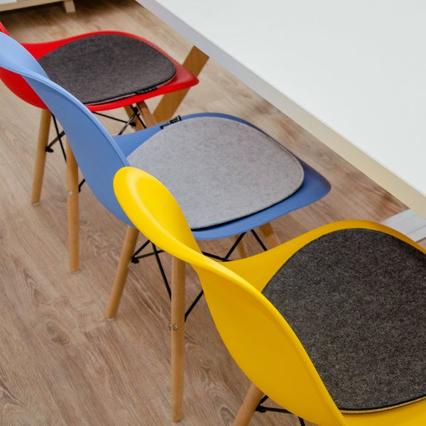 Drei moderne Schalenstühle in Rot, Blau und Gelb mit grauen Sitzkissen aus Filz und Holzbeinen, platziert an einem weißen Tisch.