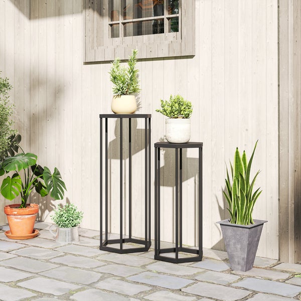 Zwei schwarze sechseckige Metall-Blumenständer unterschiedlicher Höhe mit Pflanzen auf einer hellen Terrasse vor einer Holzwand.