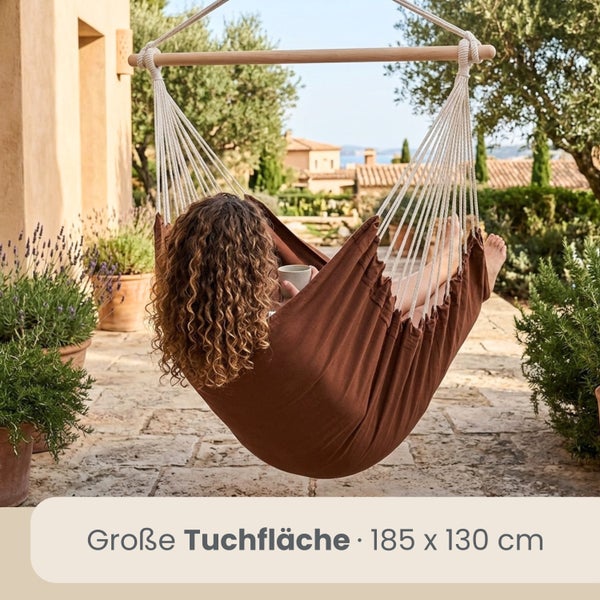Eine Person entspannt in einem braunen Hängesessel aus Stoff mit Holzstab auf einer hellen Terrasse, Tuchfläche 185 mal 130 Zentimeter.