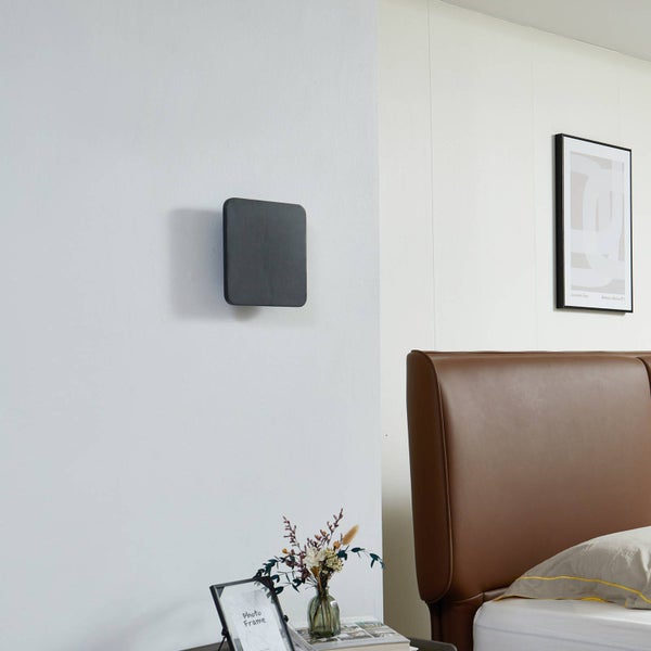 Moderne quadratische Wandleuchte in Dunkelgrau an einer weißen Wand im Schlafzimmer neben einem braunen Lederkopfteil.