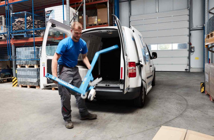 Ein Mann verlädt einen blauen Hanselifter Hubwagen in das Heck eines weißen Lieferwagens in einer Lagerhalle.