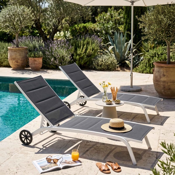 Zwei moderne Gartenliegen aus weißem Aluminium mit grauer Polsterung am Pool, umgeben von mediterranen Pflanzen und Gartenmöbeln.