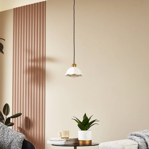 Moderne Pendelleuchte mit weißem Keramikschirm, gewelltem Rand und goldfarbener Fassung an schwarzem Textilkabel in hellem Wohnraum.