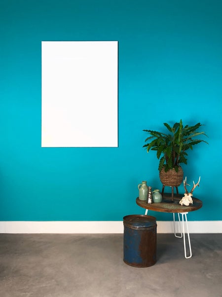 Türkisfarbene Wandgestaltung mit leerem weißem Posterrahmen. Kleiner runder Beistelltisch mit Pflanze und Dekorationsartikeln auf grauem Boden.