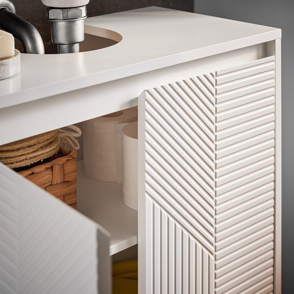 Detailansicht eines weißen Waschtischunterschranks mit geometrischem Design an den Türen, inklusive Stauraum mit Korb und Toilettenpapier