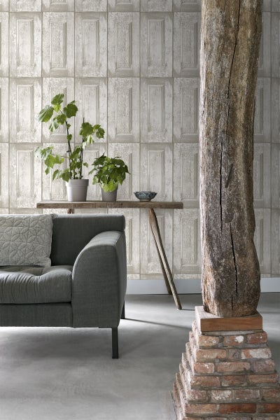 Wohnzimmer mit grauer Couch, Holztisch, Zimmerpflanzen und Holzpfosten auf Ziegelsteinsockel vor einer Wand mit Paneeltapete