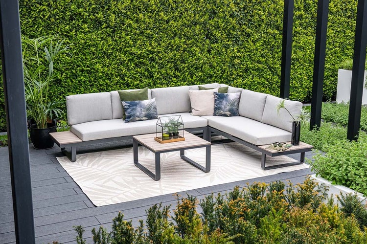Moderne Gartenlounge aus Metall mit Ecksofa, hellen Polstern und quadratischem Couchtisch auf einer Terrasse vor einer grünen Hecke.