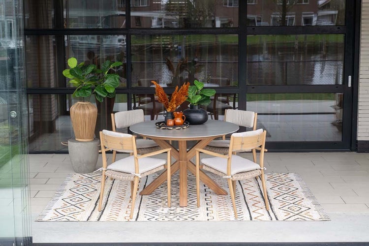 Moderne Terrasse mit rundem Gartentisch aus Holz und Steinoptik-Platte, vier Holzstühlen mit hellen Polstern auf einem gemusterten Outdoor-Teppich.