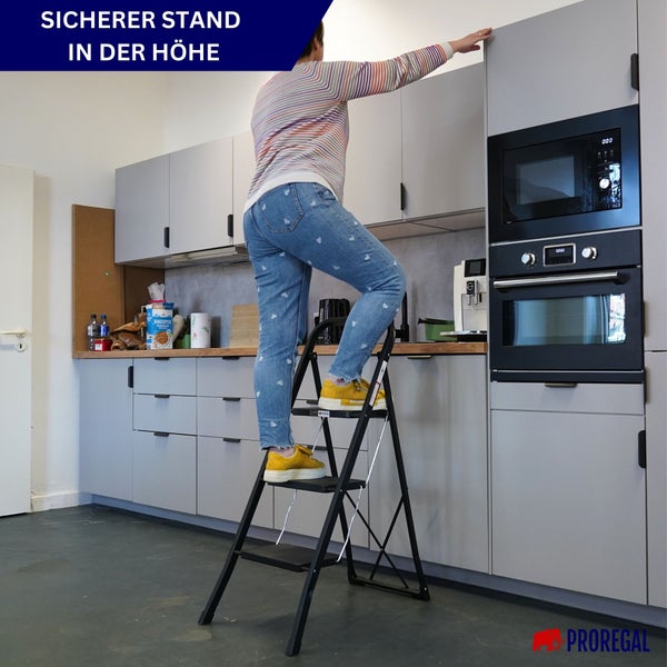 Eine Person nutzt eine schwarze Klapptrittleiter mit drei Stufen in einer Küche, um einen Oberschrank zu erreichen. Proregal Logo.