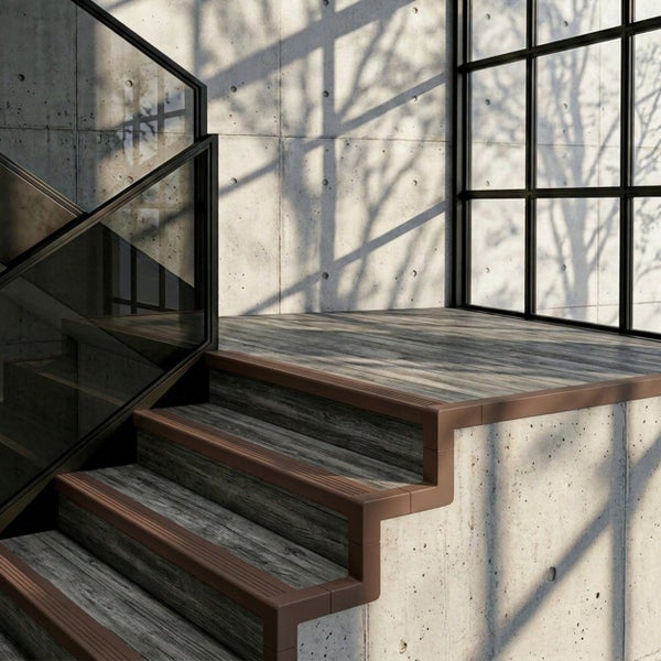 Moderne Treppe mit braunen Stufenkantenprofilen, Stufen in Holzoptik, Glasgeländer und Sichtbetonwänden in hellem, industriellem Ambiente.