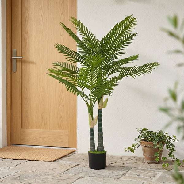 Künstliche Palmenpflanze mit zwei Stämmen im schwarzen Topf auf einer Steinterrasse neben einer Holztür und einer Efeupflanze.
