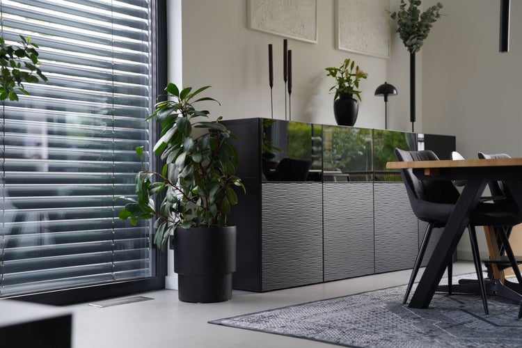 Helles, organisiertes Wohnzimmer mit schwarzem Sideboard, Jalousien am großen Fenster, Zimmerpflanze im Topf und Esstisch mit Stuhl.