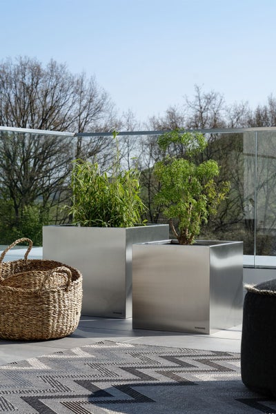 Zwei quadratische Pflanzkübel aus Edelstahl mit Pflanzen auf einer Terrasse neben einem Weidenkorb und einem gemusterten Outdoor-Teppich.