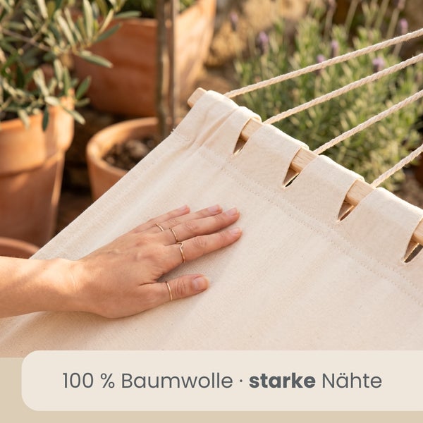 Eine Hand berührt den Stoff einer Hängematte aus 100 Prozent Baumwolle mit starken Nähten und Spreizstab aus Holz in einem hellen Garten.