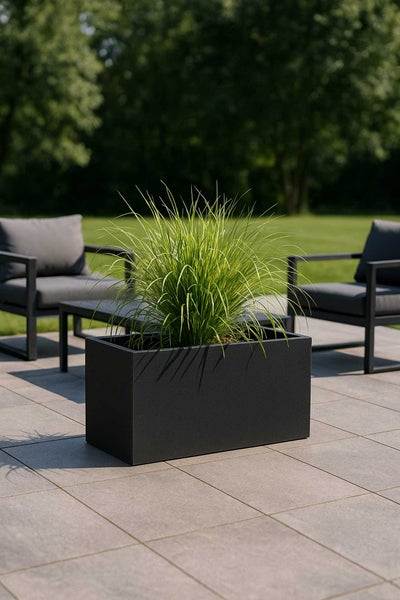 Rechteckiger Pflanztrog in Dunkelgrau mit Ziergras auf einer gefliesten Terrasse, im Hintergrund moderne Gartenmöbel und Rasen.
