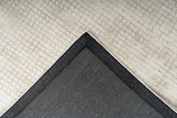Detailansicht eines Teppichs mit hellbeiger, strukturierter Oberfläche und schwarzer, rutschhemmender Unterseite an der Ecke.