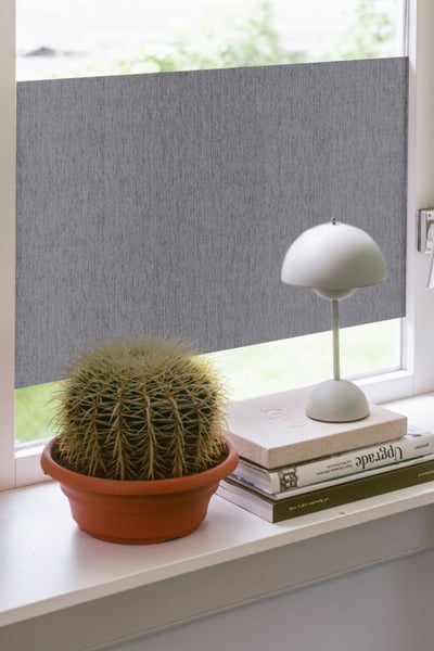 Graues strukturiertes Rollo an einem Fenster mit Kaktus und kleiner Lampe auf der Fensterbank.