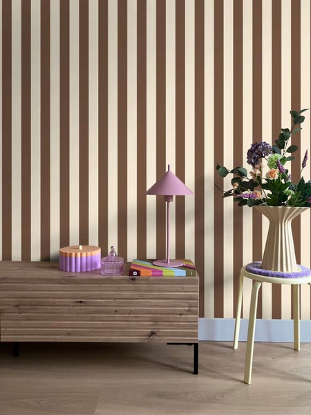 Wohnzimmer mit Vliestapete im vertikalen Streifendesign, kombiniert mit einem Holzsideboard, einer Tischleuchte und einer Vase mit Blumen.