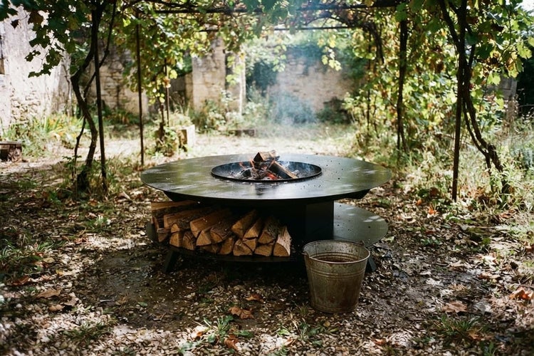 Runde Feuerschale mit Grillplatte und integriertem Holzfach im Garten unter einer Weinlaube mit brennendem Holzfeuer.