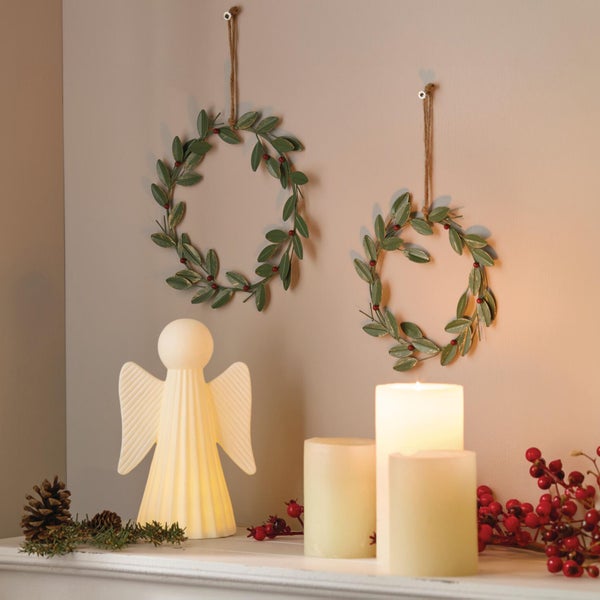 Weihnachtliche Dekoration mit zwei hängenden Metallkränzen, einer leuchtenden Engelsfigur und Stumpenkerzen auf einem Kaminsims.