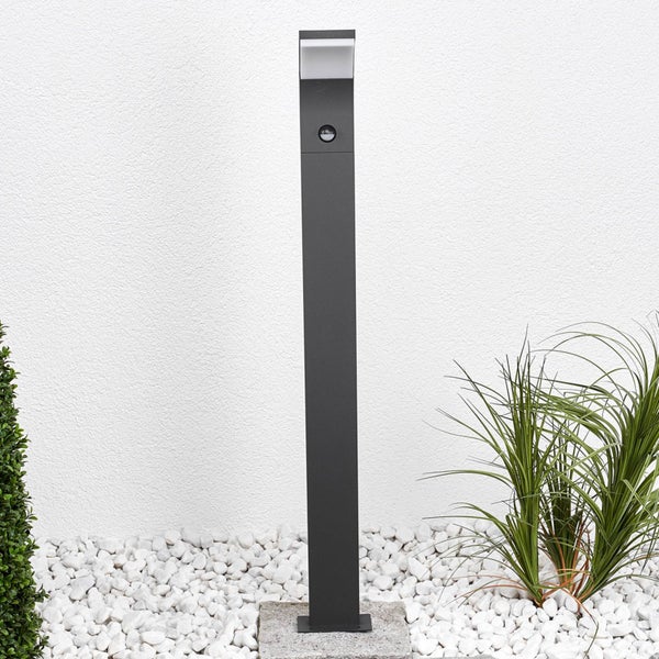 Moderne anthrazitfarbene LED-Pollerleuchte mit Bewegungsmelder in einem Garten mit weißem Kies und Pflanzen.