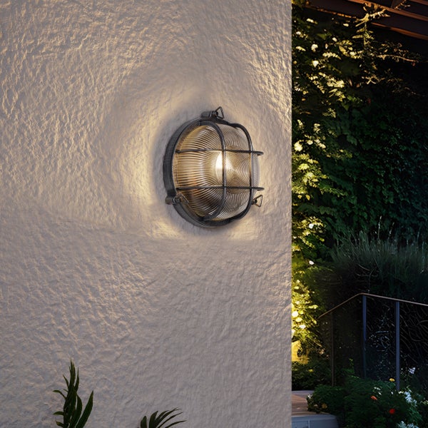 Beleuchtete runde Außenwandleuchte mit Metallkäfig und geripptem Glas, montiert an einer strukturierten Wand in einem Garten.