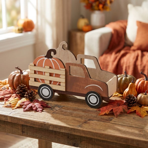 Dekorativer Holz LKW mit Kürbis für den Herbst auf einem rustikalen Holztisch umgeben von herbstlicher Dekoration wie Zapfen und künstlichen Blättern.