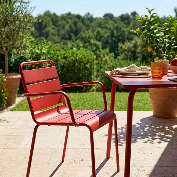Roter Gartenstuhl aus Metall mit Armlehnen und Lattenrost, passender roter Gartentisch auf einer sonnigen Terrasse mit Pflanzen im Hintergrund.