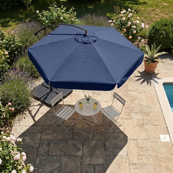 Ein runder blauer Ampelschirm über einer weißen Bistro-Sitzgruppe aus Metall auf einer Naturstein-Terrasse im Garten neben einem Pool.