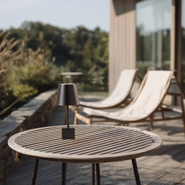 Dunkle Akku-Tischleuchte auf einem runden Holztisch auf einer hellen Terrasse mit Sonnenliegen im Hintergrund.